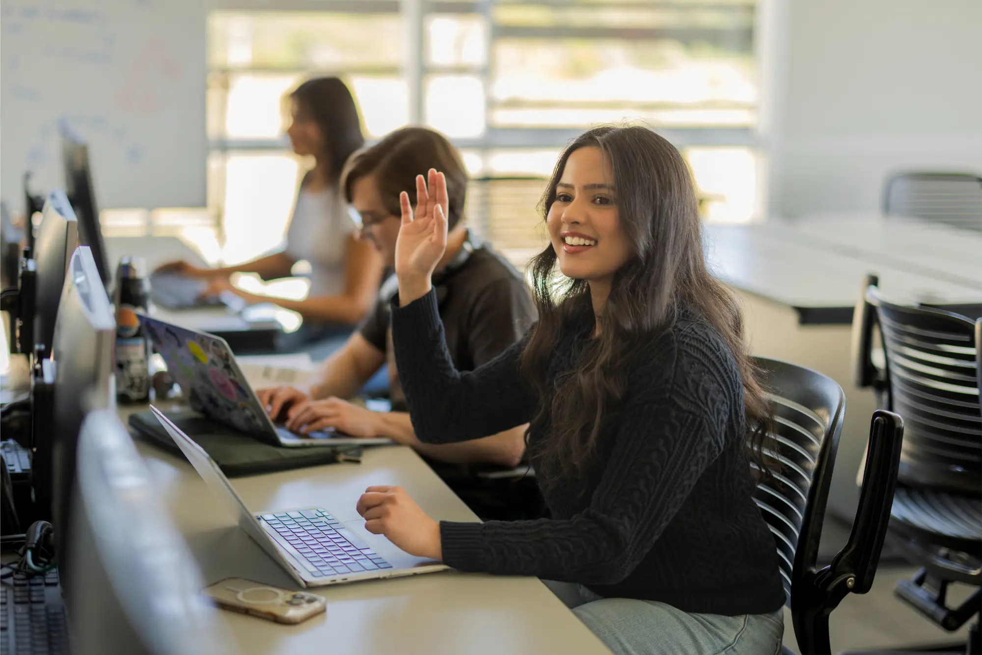 A group of students working while one has their hand raised asking a question