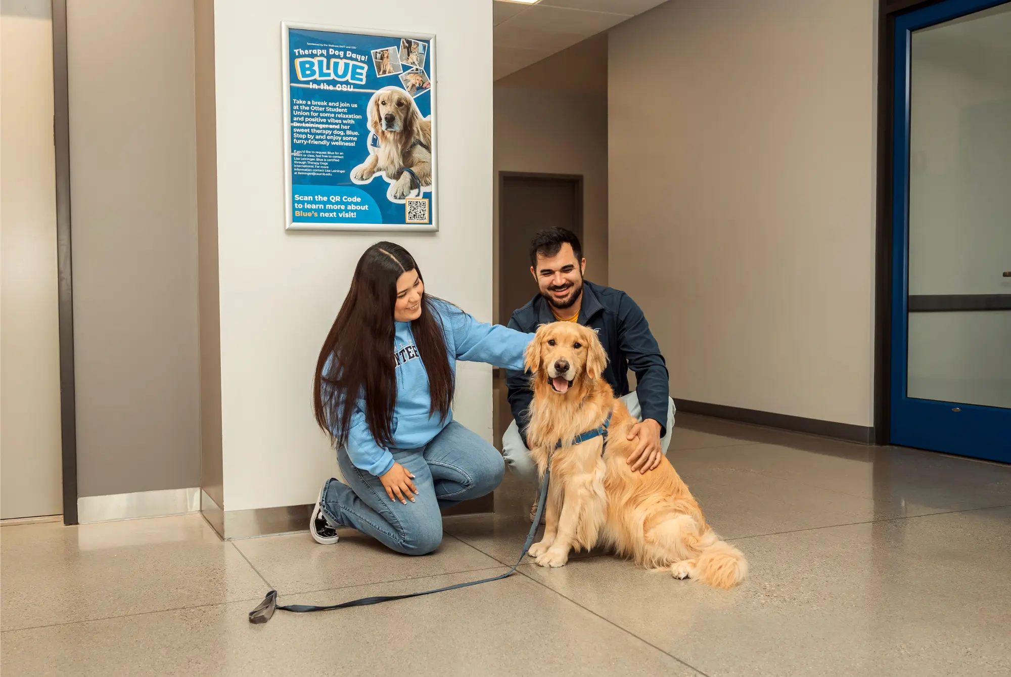 Two Students In front of the elevator inside the OSU one male one female petting a golden retriever therapy dog named Blue.