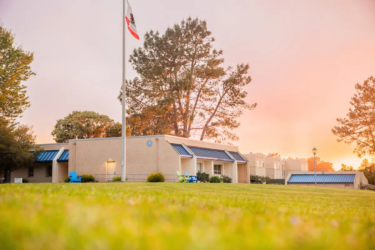 A campus lawn glows at sunset with buildings, trees, and U.S. and a California flag under a sky filled with golden clouds.