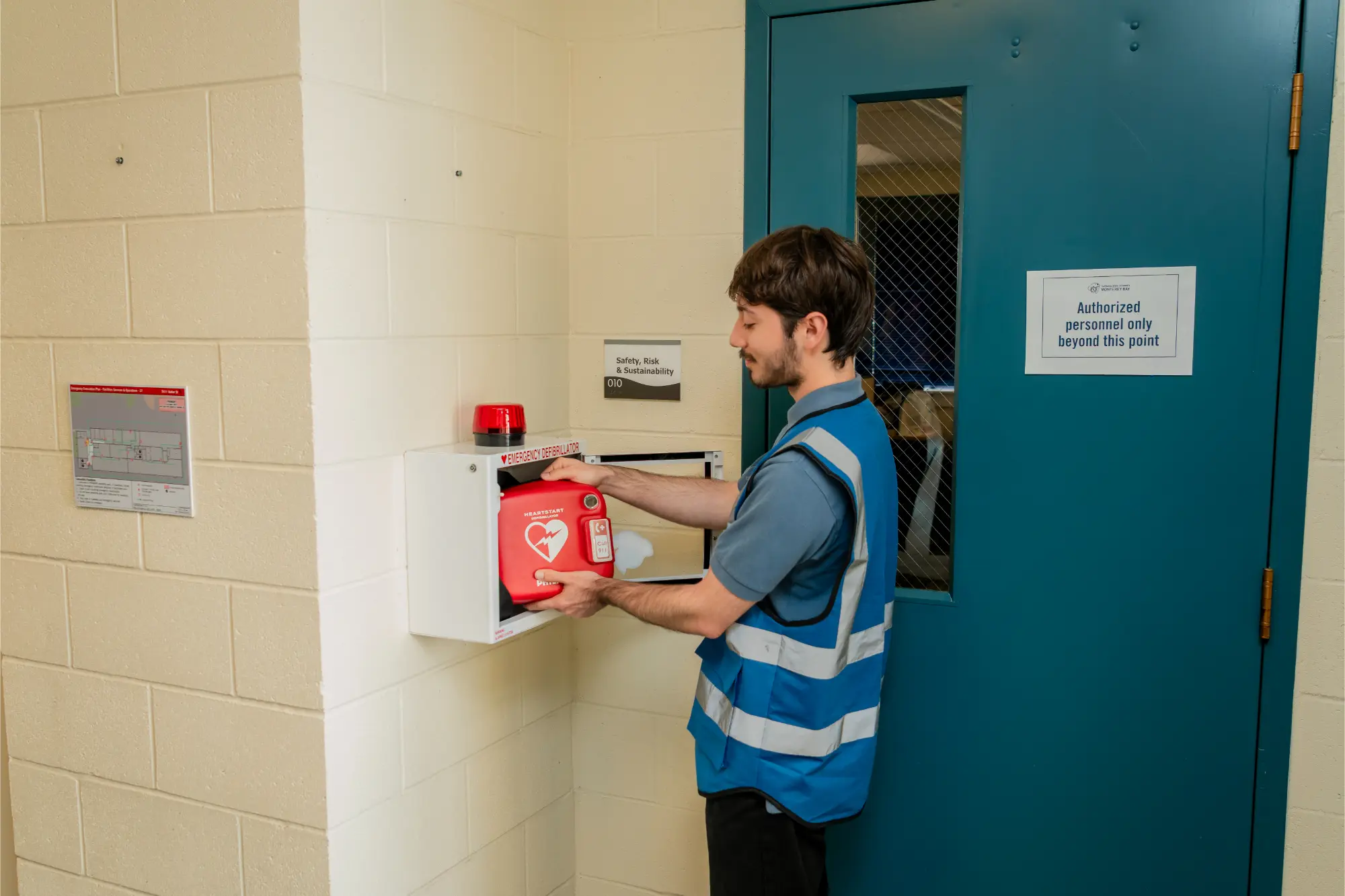 The same student retrieves an AED (defibrillator) from a wall-mounted emergency case outside the Safety, Risk & Sustainability office door.