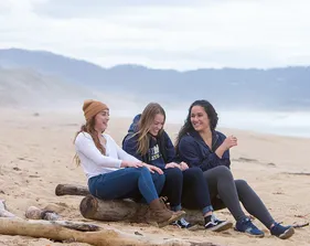 Three students sitting on a log on the beach laughing.