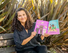 A woman sitting on a bench outdoors smiles while holding open a colorful children’s book featuring illustrations of raised fists and people eating tacos, with text in both English and Spanish.