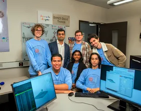 Three students sitting and smiling for a photo with three students standing behind them and the professor also smiling