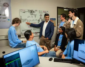 Six students looking at the professor teach while he is pointing at a whiteboard.