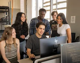A group of students looking at a students desktop while they work on it