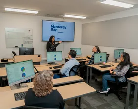 A CSUMB finance staff member presents to colleagues in a training room while standing in front of a display reading 