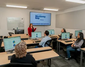 A staff trainer at CSUMB presents a Procurement and Contracts session in a finance department training room as staff listen attentively from their computers.