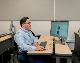 A CSUMB finance staff member reviews colorful financial pie charts on a desktop monitor, focusing intently while seated at a classroom workstation.