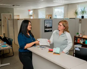 A CSUMB HR representative smiles while handing an employee paperwork across a front desk in a bright and welcoming office, decorated with plants and inspiration posters.