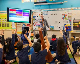 A teacher stands at the front of an elementary school classroom, pointing toward students who are seated on the floor with their hands raised.