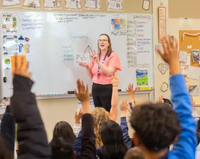 Teacher stands at the whiteboard while students raise their hands in class
