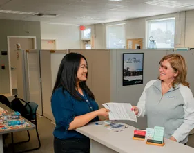 A ���ӽ紫ý HR representative smiles while handing an employee paperwork across a front desk in a bright and welcoming office, decorated with plants and inspiration posters.
