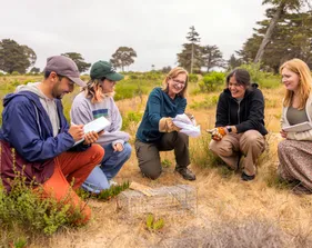 A group of four AES students and a professor kneeling in the grass while the professor holds a squirrel