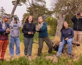 A group of five AES students and a professor standing in the grass using binoculars
