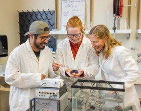Students in lab coats observe and point to a tank experiment as a professor explains the setup in a marine science lab