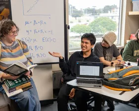 Students smiling while discussing a math topic written on the whiteboard next to them.