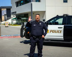A male CSUMB UPD  officer smiles and waves in front of the police SUV. Campus buildings and a clear sky complete the friendly scene.