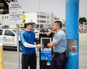 A CSUMB parking officer chats with a student at a parking kiosk. A “Parking Information” sign and a Parking Services truck are in the background.