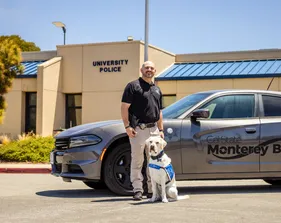A CSUMB officer stands smiling beside the seated service dog in front of a Monterey Bay police car and the University Police station.