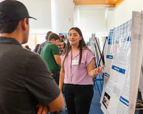 A student presenter points to her graphic board while presenting to an onlooker in the audience