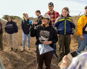 A group of students stands in a circle with a women in the middle holding up a field mouse.