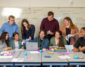 Students collaborating with each other in front of a desk, some of them sitting and some standing.