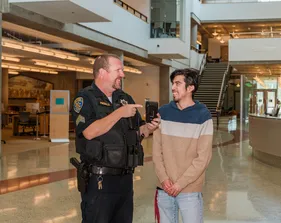 A ���ӽ紫ý sergeant laughs with a student inside the library, pointing to a device in his hand during a friendly conversation.