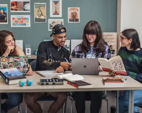 A group of four students sitting at a table collaborating with computers, books, and papers on the table.