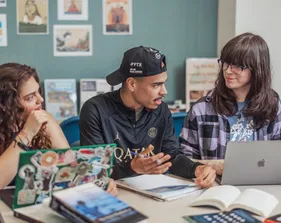 A group of three students talking together while they have computers and papers out on the table.