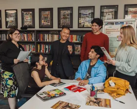 A group of students standing and sitting smiling while a professor stands in the middle smiling pointing at the camera.