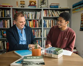 A student and professor meeting with each other smiling and looking over some papers.