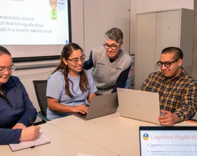 Three professors sitting at a table on their computer and taking notes while discussing with a student also sitting at the table