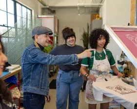 A student printing a design on a shirt using paint and a professor helping her out with multiple people in the background.