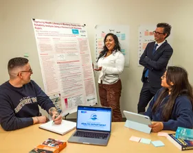 Student presenting a poster of work to two other students sitting at a table listening and a professor standing behind the presenter, also listening.