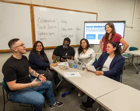 Five students sitting at a table listening while one of them is speaking to the professor who is standing smiling