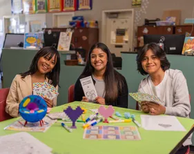 A social work credential student and two kids sitting at a desk in a classroom while smiling at the camera