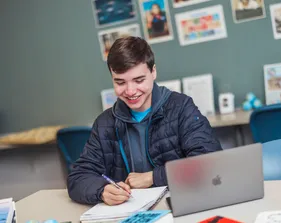 A student doing work on paper while having a computer open and smiling