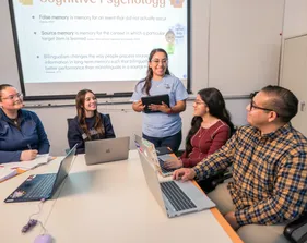 Multiple students and professors sitting at a table listening to a girl presenting to them.