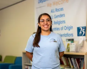 Student standing with her arms behind her back smiling for a portrait inside.
