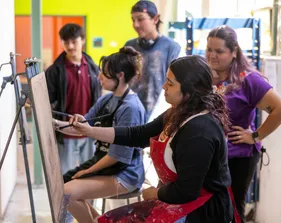 Three students standing and smiling around two students who are sitting and painting on canvases.