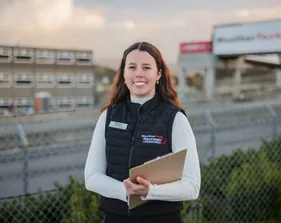 A student smiling for a portrait in front of the race track while holding a clip board.