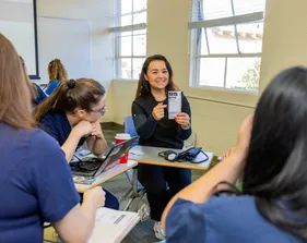 Student presenting something to her classmates while sitting at a table in a classroom.
