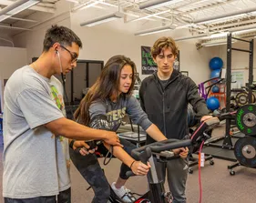 A student using gym equipment with two other students assisting.