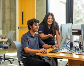 A smiling student sitting down at a desk using a computer, with a school counselor peering over their shoulder and pointing at the computer screen with a smile