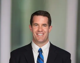 Portrait of Josh Warburg smiling in a dark suit and blue striped tie against a soft green background.