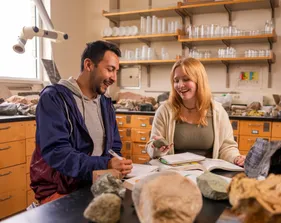 Two people are seated in a laboratory. One holds a stone in her hand, while the other uses a pen to write
