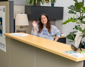 A woman stands behind a desk, smiling warmly and waving hello.