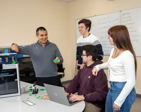 A student sitting in front of laptop and three people standing around him