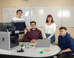 A group of four mechatronics students smiling at the camera next to a 3d printer