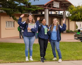 Three female students wearing CSUMB gear, one holding a clipboard, smile and wave at the camera.